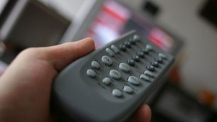 Hand holds a gray remote control pointing at a blurry TV screen. The background is indoors, creating a relaxed, focused mood.