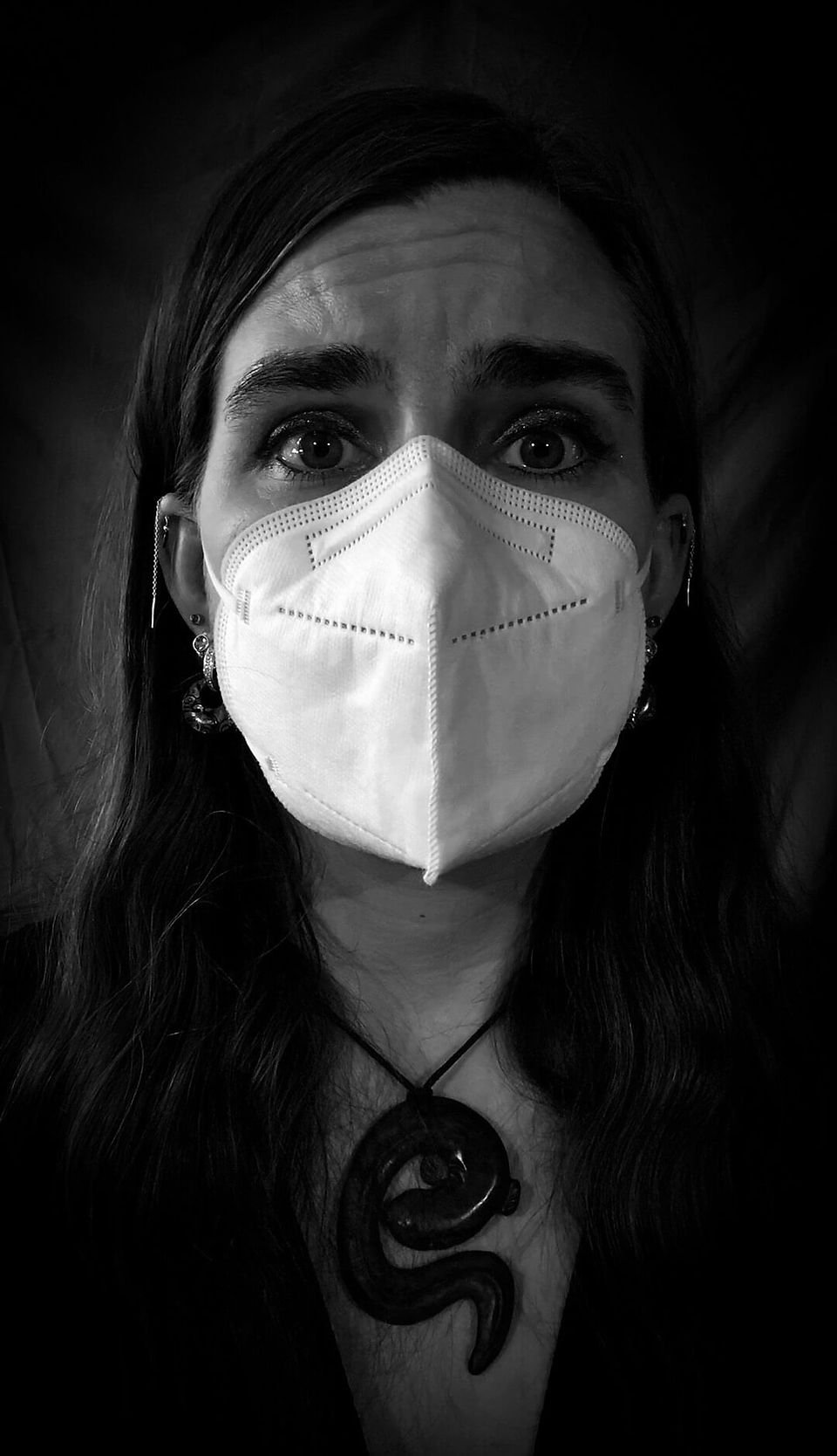 Person wears a white face mask, looks worried. Black and white setting, with a dark background. Long hair and ornate necklace visible.