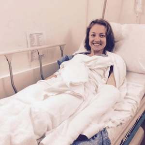 Smiling woman in hospital bed, wrapped in white blanket, wearing a gown. Hospital room setting with beige walls.