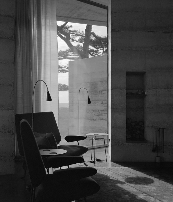 Black and white photo of a cozy corner inside the Secular Retreat by Peter Zumthor, showcasing modern chairs and floor lamps with natural light streaming in.