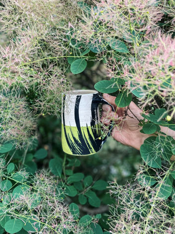 A black, white and chartreuse beer stein being held by hands partially concealed by smoke bush leaves, by Whitney Gill, 2025