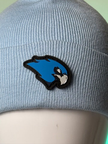 A light blue knit beanie with a blue and black bird logo displayed against a blurred light background.