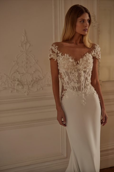 A crepe and lace sheath style wedding dress with a sheer bodice, cap sleeves, low back and long train. It is also available with an additional detachable train for added drama.
