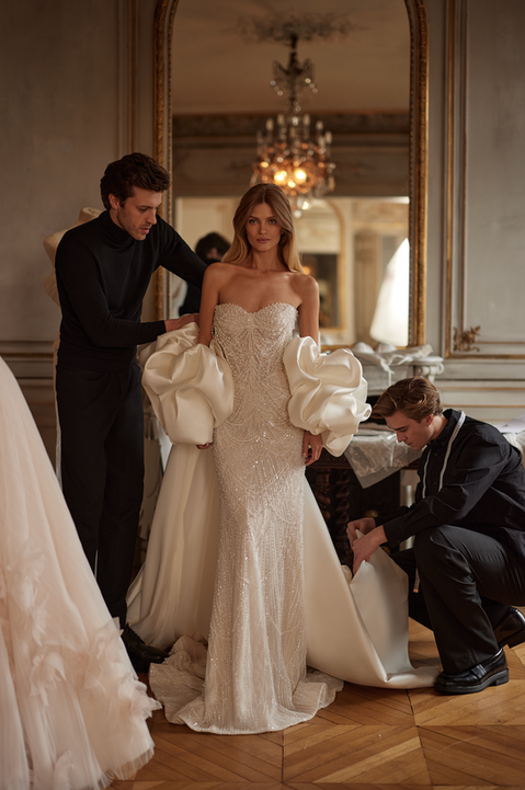 A strapless sheath wedding dress richly decorated with beads. Also available with a dramatic puff sleeve cape.