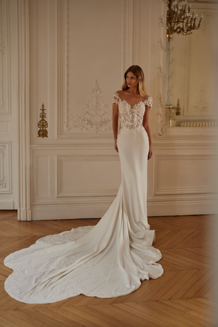 A crepe and lace sheath style wedding dress with a sheer bodice, cap sleeves, low back and long train. It is also available with an additional detachable train for added drama.