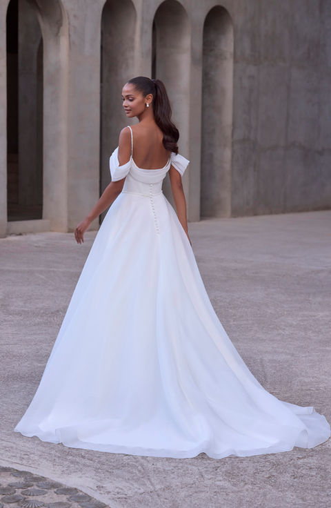 Our Helene wedding dress has a heavenly design featuring an A-line organza silhouette with a crystal and pearl embroidered sweetheart neckline framed by detachable off-the-shoulder sleeves and sparkling crystal straps. A draped organza bust and soft cowl back add sculptural dimension, while a detachable beaded waistband defines the waist. Finished with hidden pockets and a sweeping train, the gown is romantic, versatile, and effortlessly modern.
