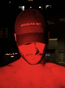 A man wearing a "Brazilian Boy" cap, illuminated by red light, stands shirtless against a dark backdrop with glowing windows