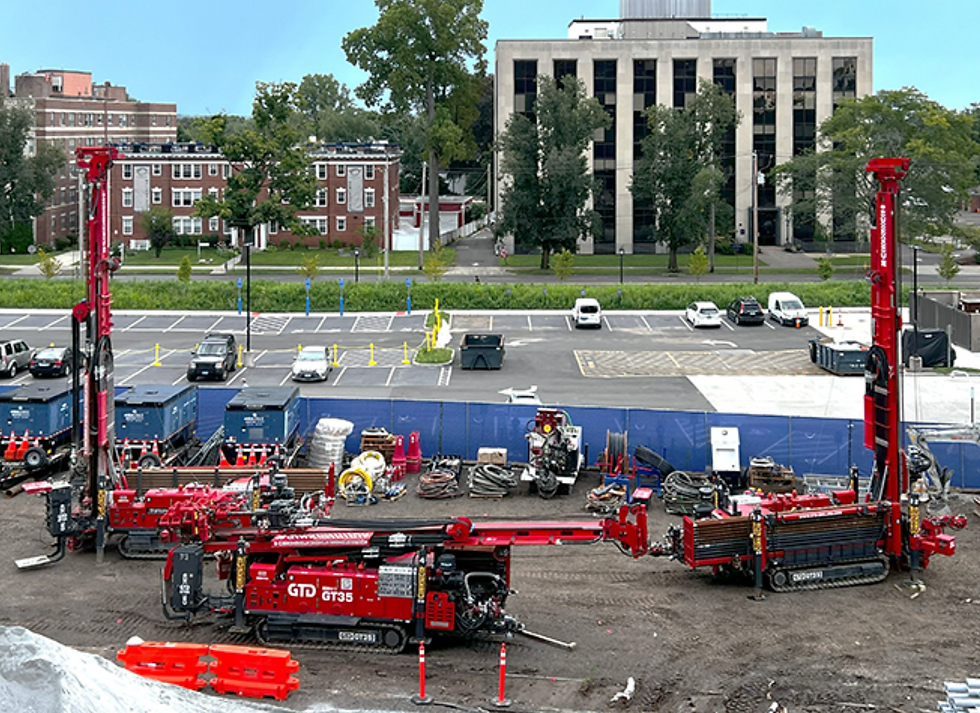 View of the parking lot on Science Hill, showing construction underway for a geothermal borefield. There are buildings in the background. Three large pieces of red equipment to perform the dig are sitting poised on a leveled dirt section of roped-off parking lot. 