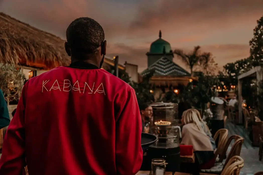 Kabana waiter serving guests at sunset on the rooftop terrace in Marrakech, with cozy lights and vibrant evening ambiance.