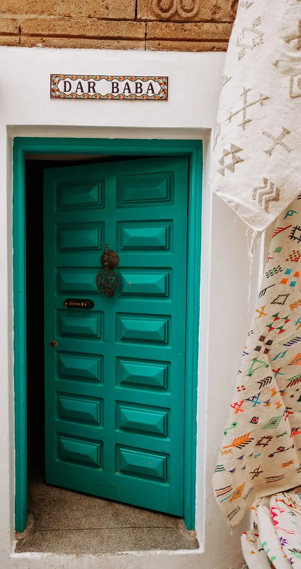 Porte d'entrée traditionnelle turquoise avec panneau Dar Baba et tapis berbères colorés dans la Médina d'Essaouira.