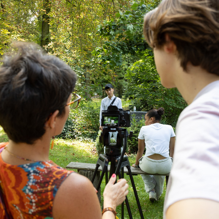 Frau und Mann filmen Szene im Park, weitere Person und Baum, Kiezbegegnung.