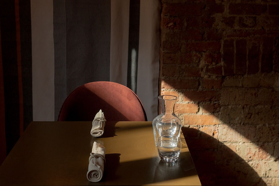 Sunlit empty restaurant table with rolled napkins, glass carafe and tumbler, red chair, and brick wall in warm shadows.