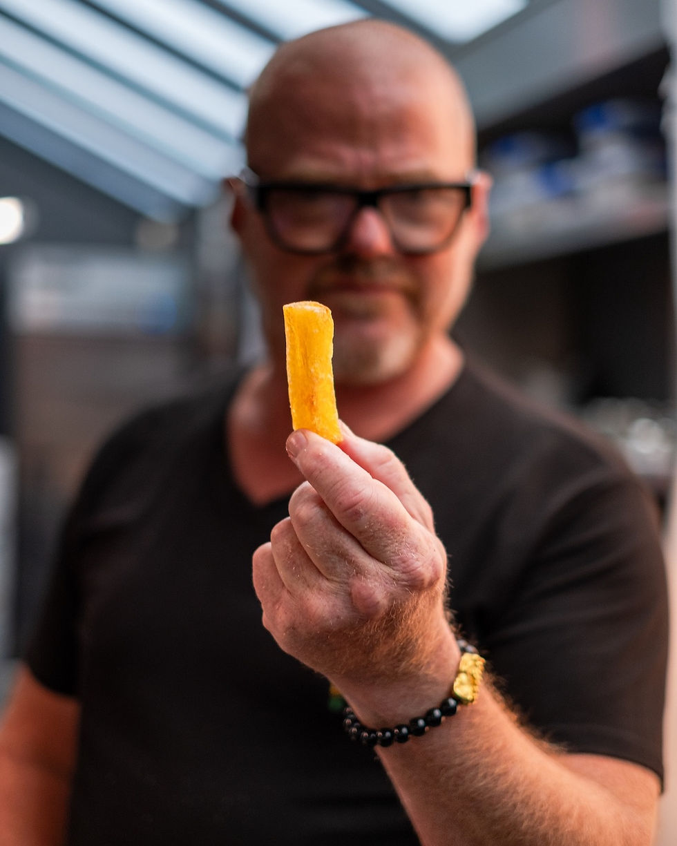 A person wearing glasses holds a golden fry, close-up. Background shows a kitchen with a striped roof. The mood is focused.