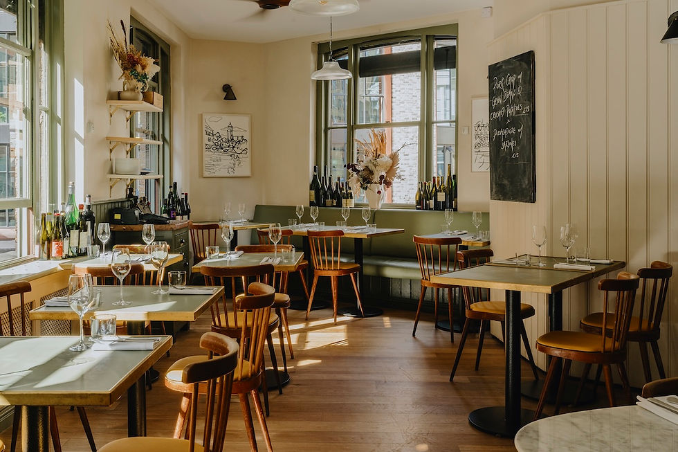 Sunny empty restaurant with set tables, wine glasses, wooden chairs, flowers, and chalkboard menu by large windows.