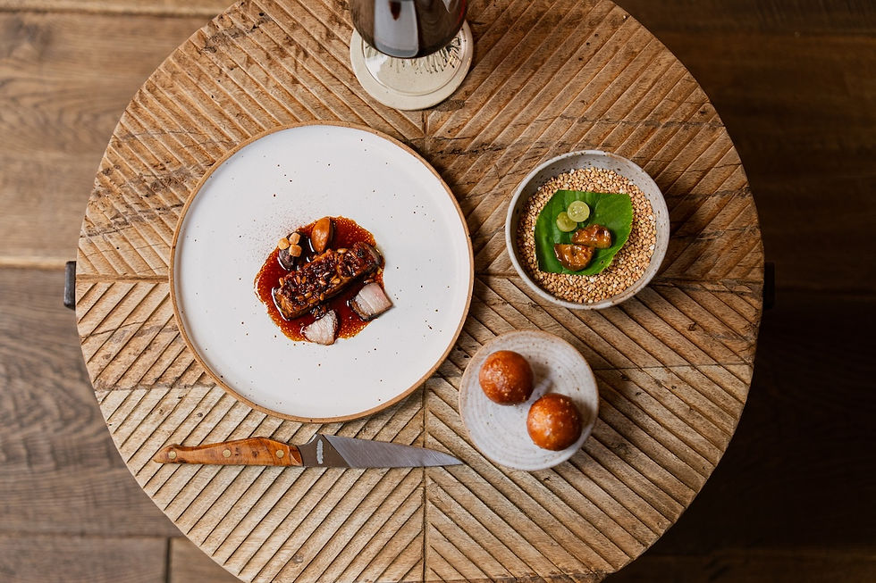 Top-down view of gourmet dishes on a round carved wooden table: plated meat, small bowls, bread rolls, and a knife.