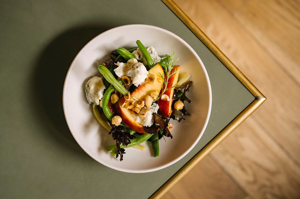 Colorful salad in a white bowl with greens, peach slices, nuts, and creamy cheese on a green table beside wood flooring