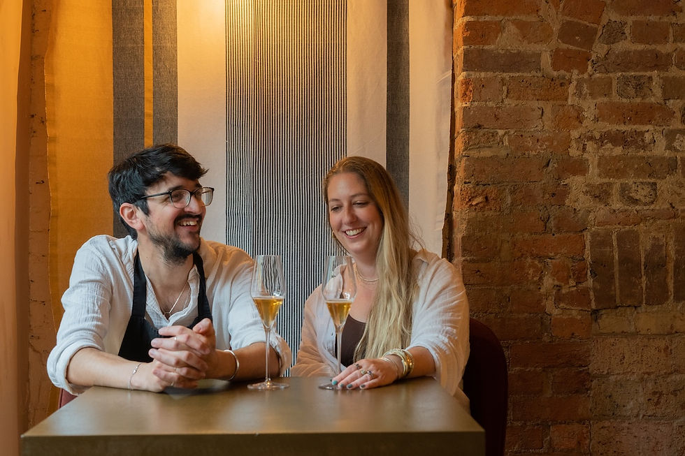 Two smiling adults sit at a table with two glasses of champagne in a cozy brick-walled bar.
