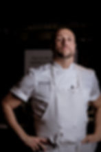 Chef in white uniform stands confidently with arms akimbo, dark background, text "BEAR by" visible on his apron.