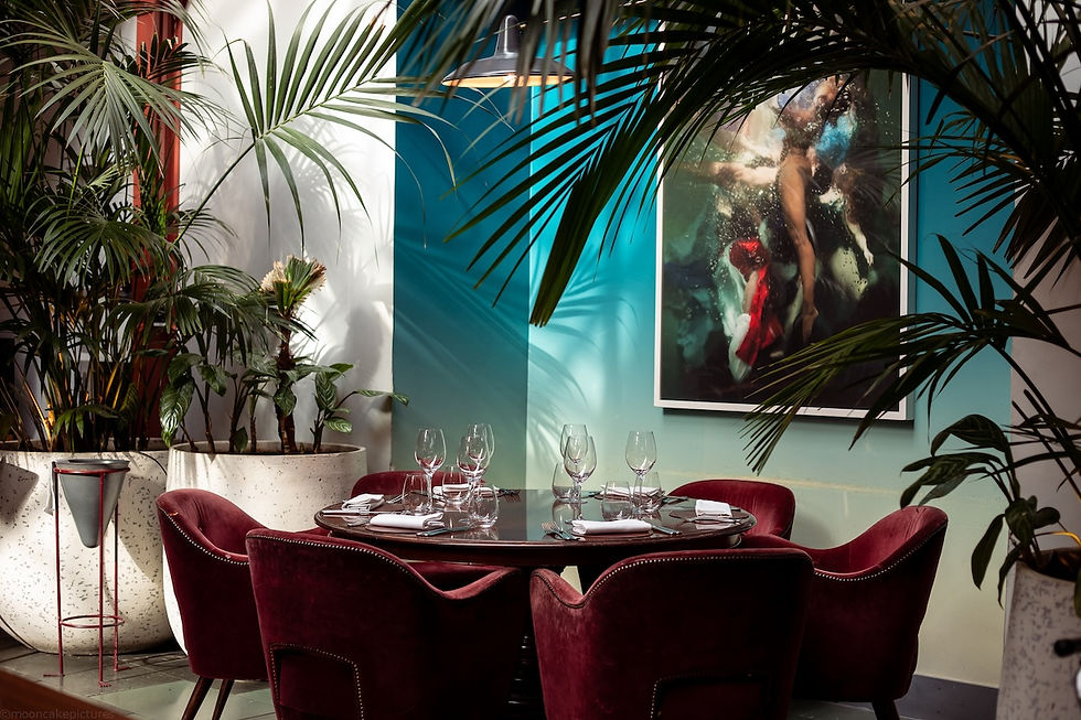 Elegant restaurant table set for four with burgundy velvet chairs, lush palms, and abstract art on a teal wall.