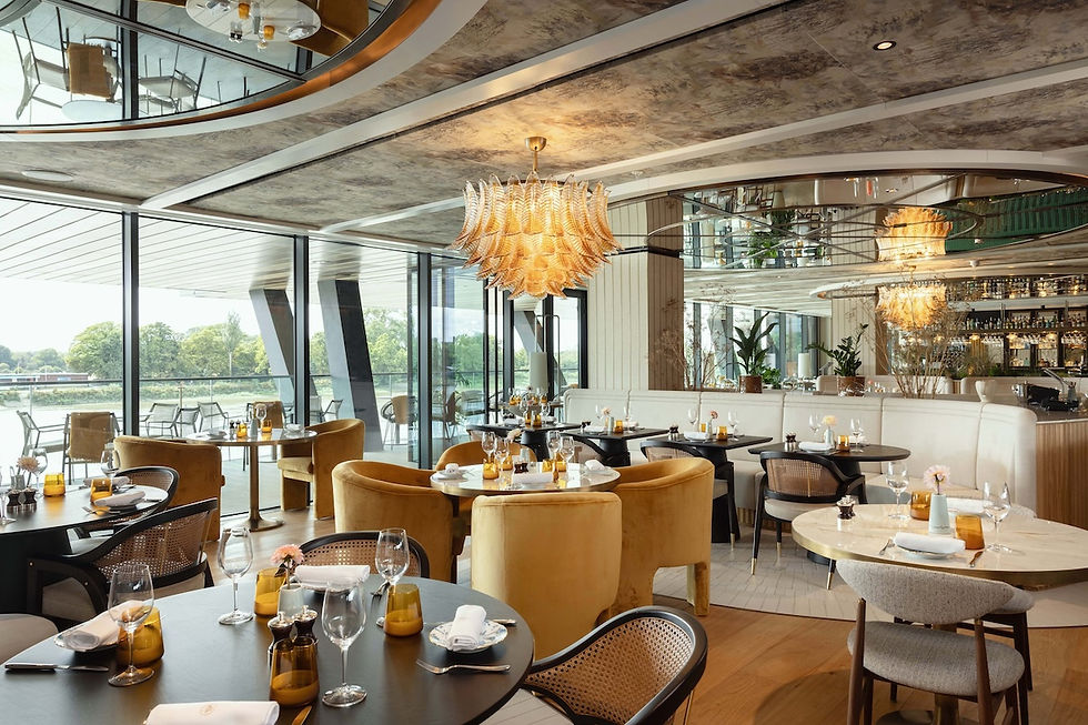 Elegant riverside restaurant with gold chandeliers, set tables, velvet chairs, and floor-to-ceiling windows overlooking water.