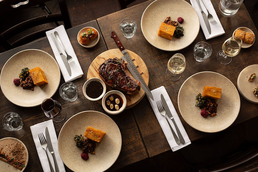 Rustic table setting with four plates, each with meat, squash, greens, and a berry. A large steak on a board with sauces. Wine and water glasses.