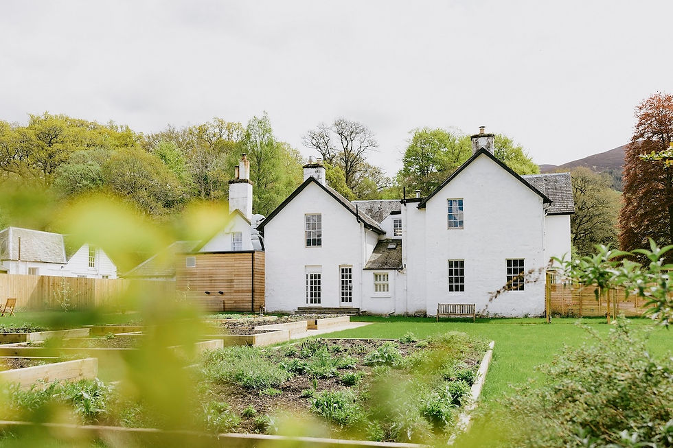 White country house with garden beds and lawn, framed by blurred green leaves, under an overcast sky and spring trees
