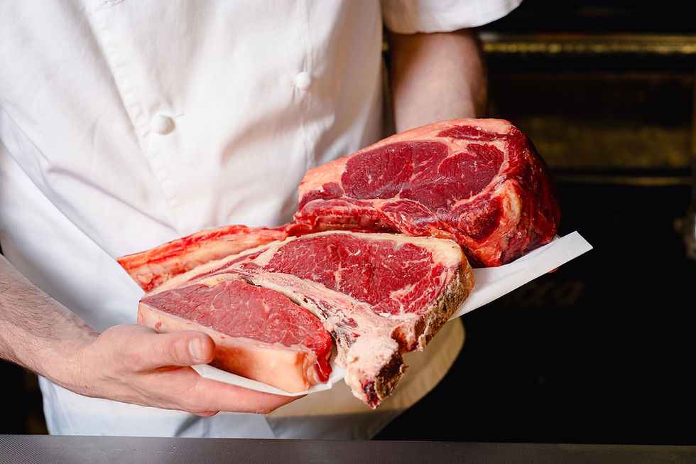 Chef in white uniform holds large raw meat cuts on paper, showcasing marbled textures. Dark blurry background highlights rich red hues.