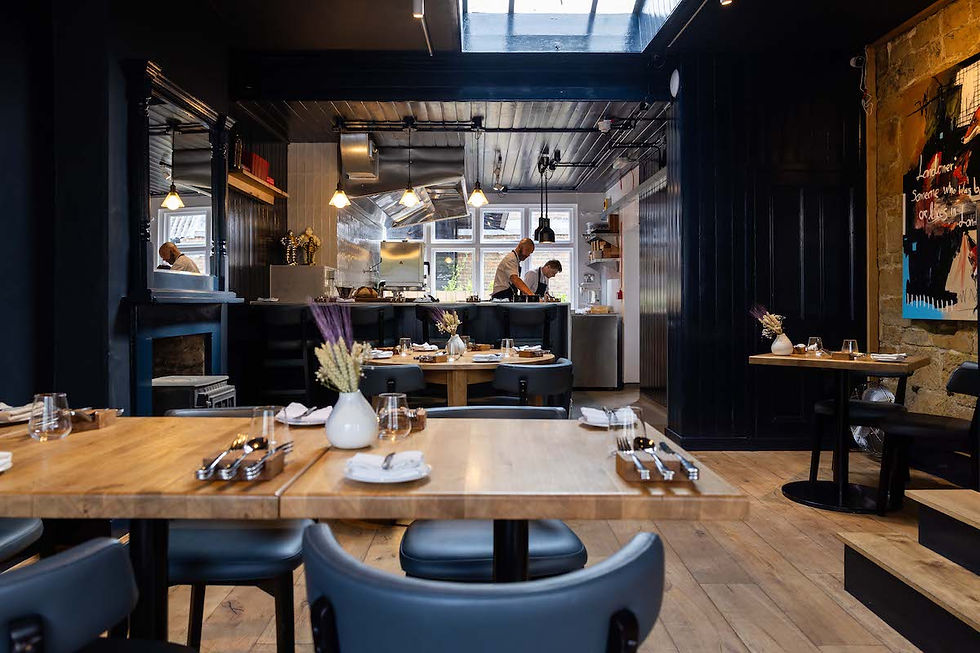 Modern restaurant dining room with set wooden tables, dark walls, and chefs working in the open kitchen.