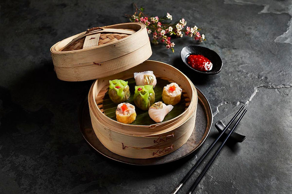 Steamed dim sum in a bamboo basket on a dark stone table. Chopsticks, red sauce, and pink flowers enhance the elegant setting.