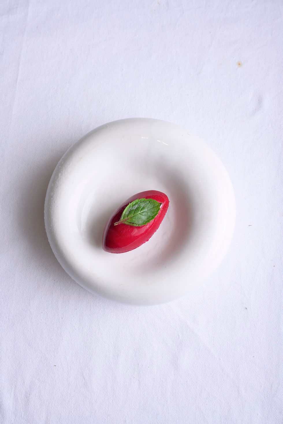 White plate with a scoop of red sorbet topped with a green mint leaf, set on a white tablecloth.