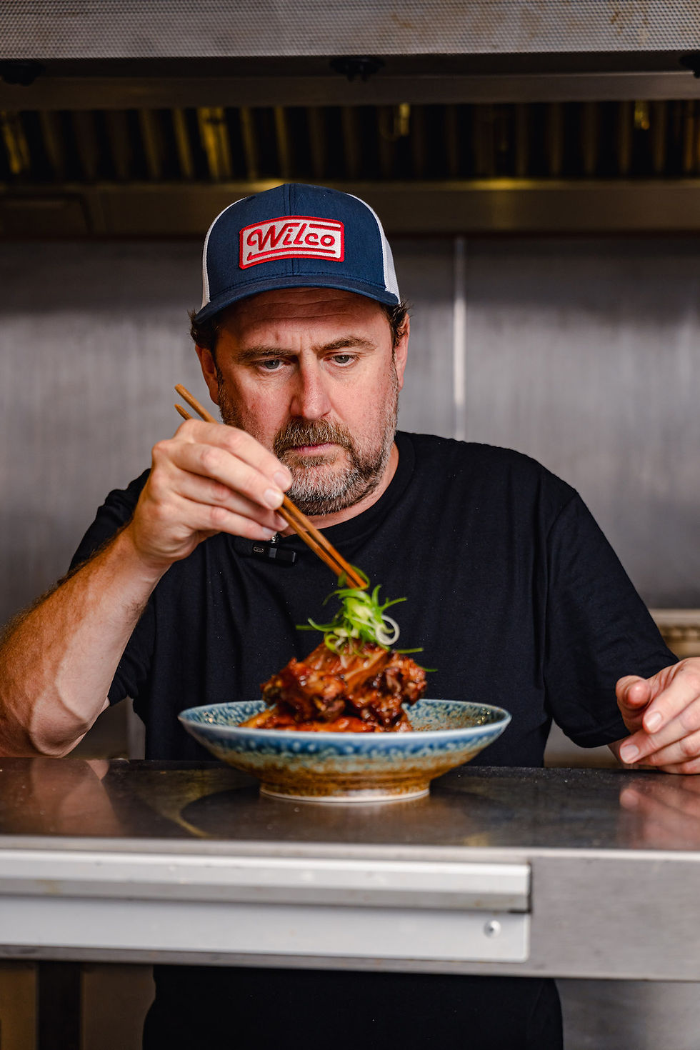 Chef in a "Wilco" cap garnishes meat dish with greens using chopsticks in a kitchen setting, focused expression, neutral background.
