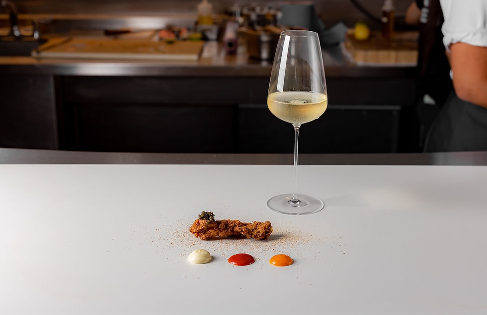 Fried chicken with caviar and three sauces on white surface, wine glass nearby. Blurred kitchen background, warm lighting.