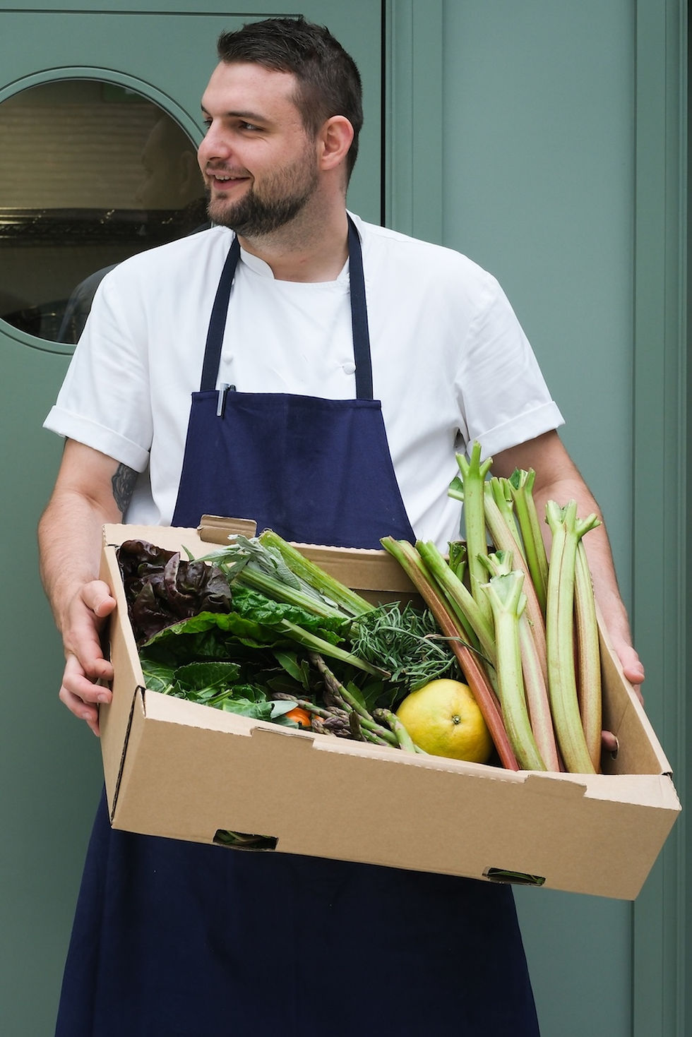 Smiling chef in a navy apron holds a box of fresh vegetables and rhubarb in front of a green door.