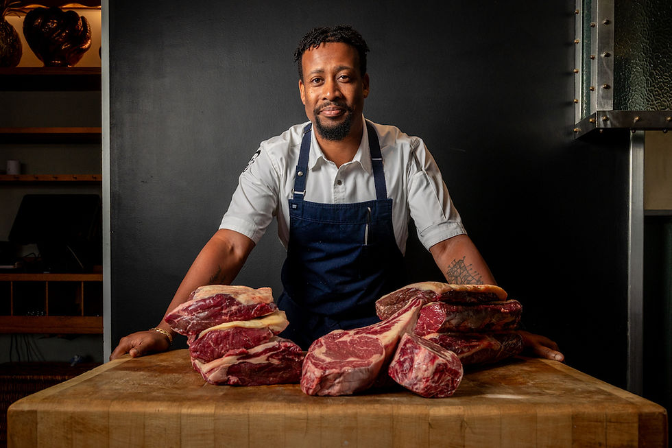 A chef in a white shirt and blue apron stands behind a wooden table with raw meat cuts, set against a dark background, looking content.