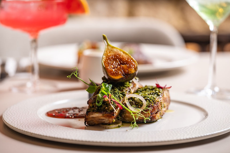 Gourmet dish with herb-crusted meat, fig, and greens on a white plate. Vibrant drinks in the background; elegant dining setting.