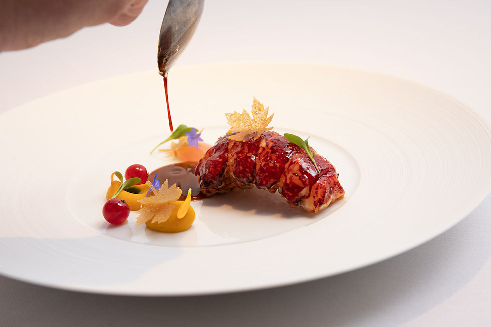 Gourmet dish with lobster tail on a white plate, garnished with colorful flowers and puree. Sauce being artistically drizzled. Elegant presentation.