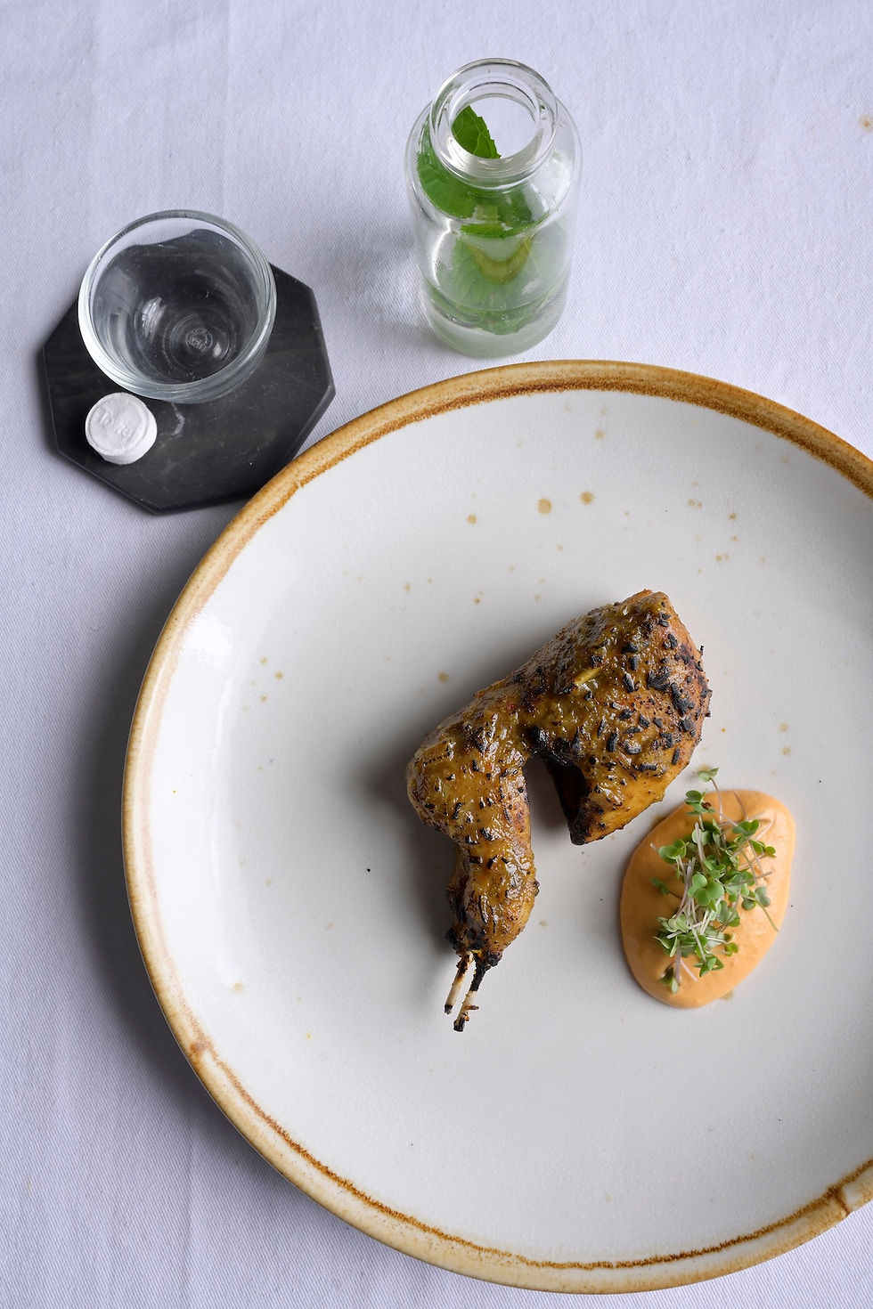 Grilled chicken wing with herbs and aioli on a white plate. Side jar with leaves, an empty glass, and a white tablecloth background.
