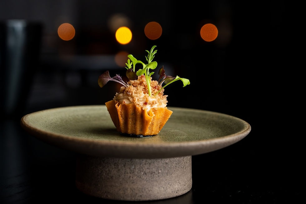 Gourmet bite in a fluted tart shell topped with greens on a stone pedestal plate, against a dark background with warm bokeh lights
