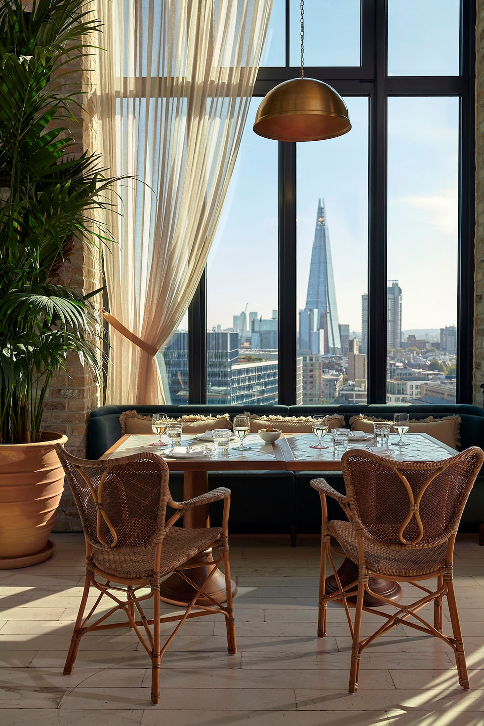 Elegant restaurant table for two by tall windows, with wicker chairs, sheer curtains, and The Shard skyline beyond