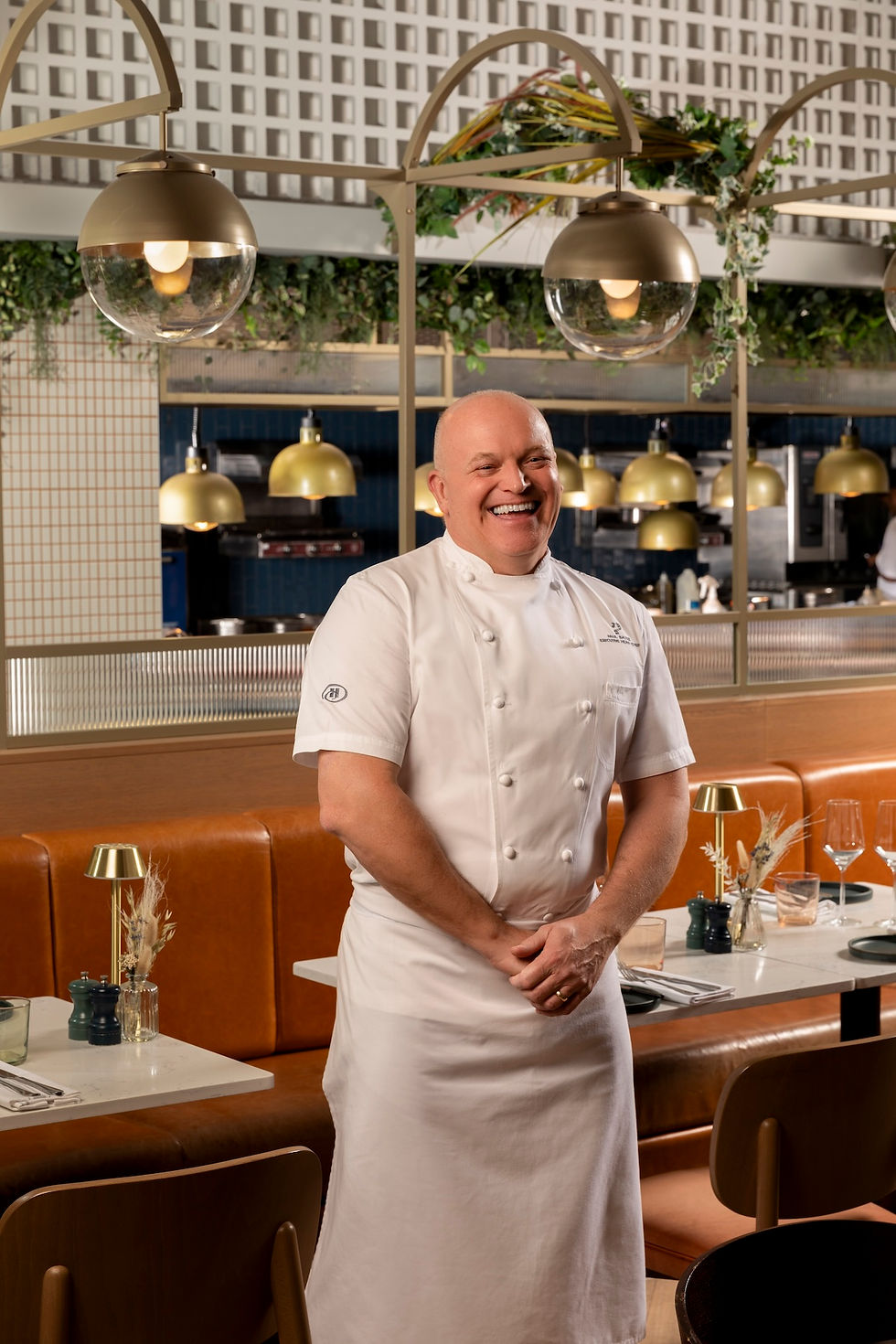 Smiling chef in white coat stands in a stylish restaurant with gold pendant lights and tan booths.