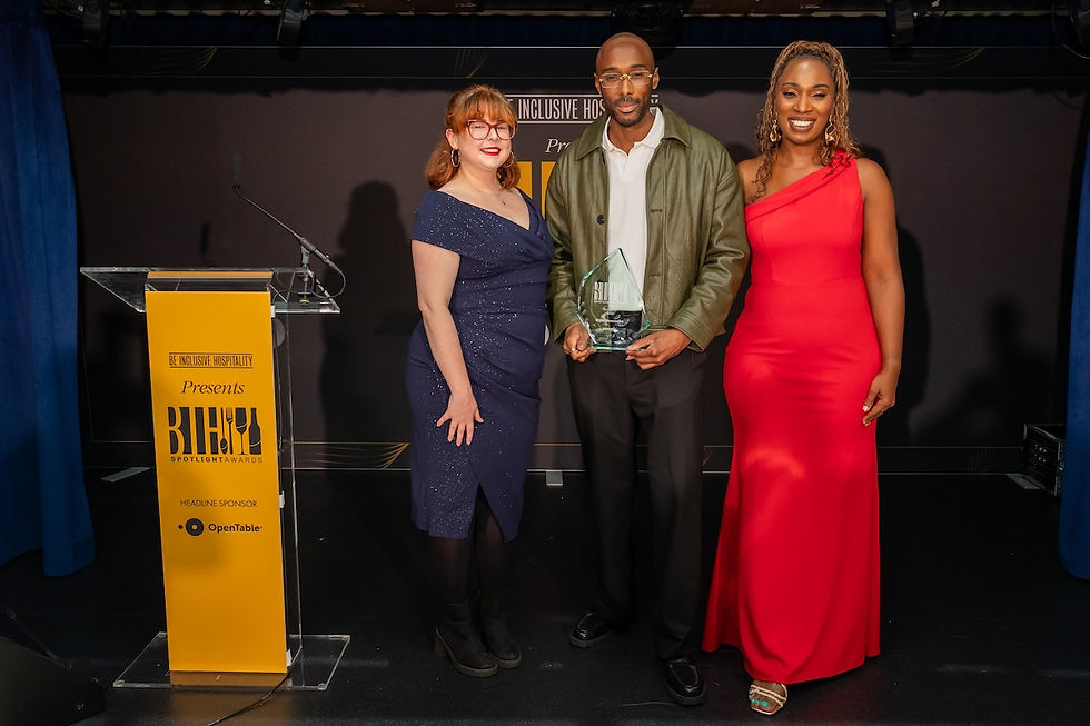 Three smiling people on stage at the Be Inclusive Hospitality Spotlight Awards, with the center man holding a glass trophy.