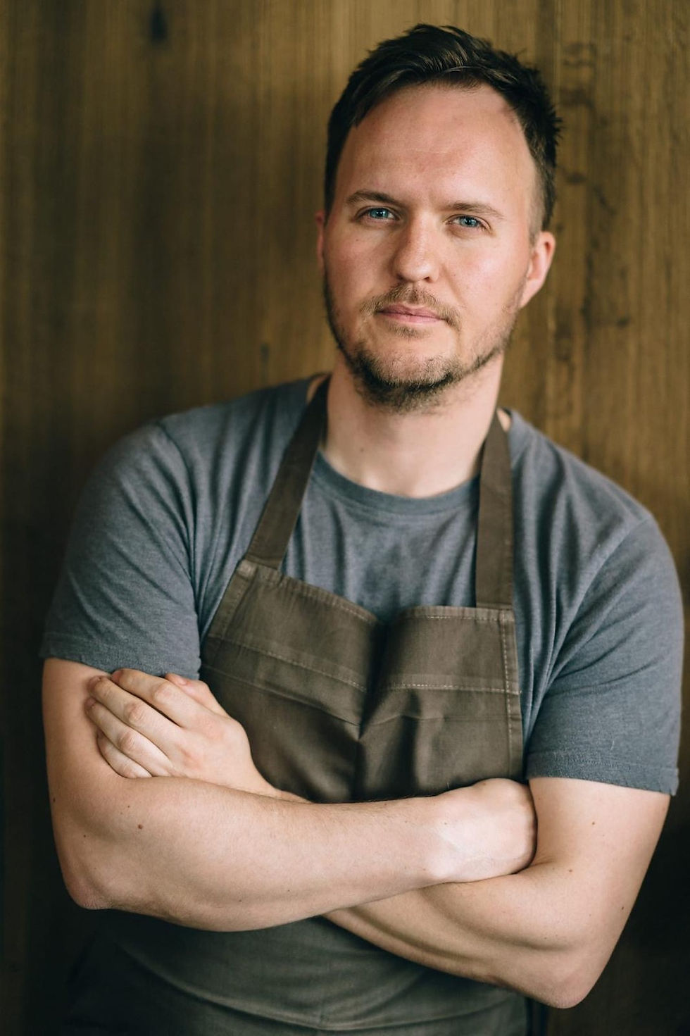 Man in a gray t-shirt and brown apron, arms crossed, standing against a wooden background. Calm expression.