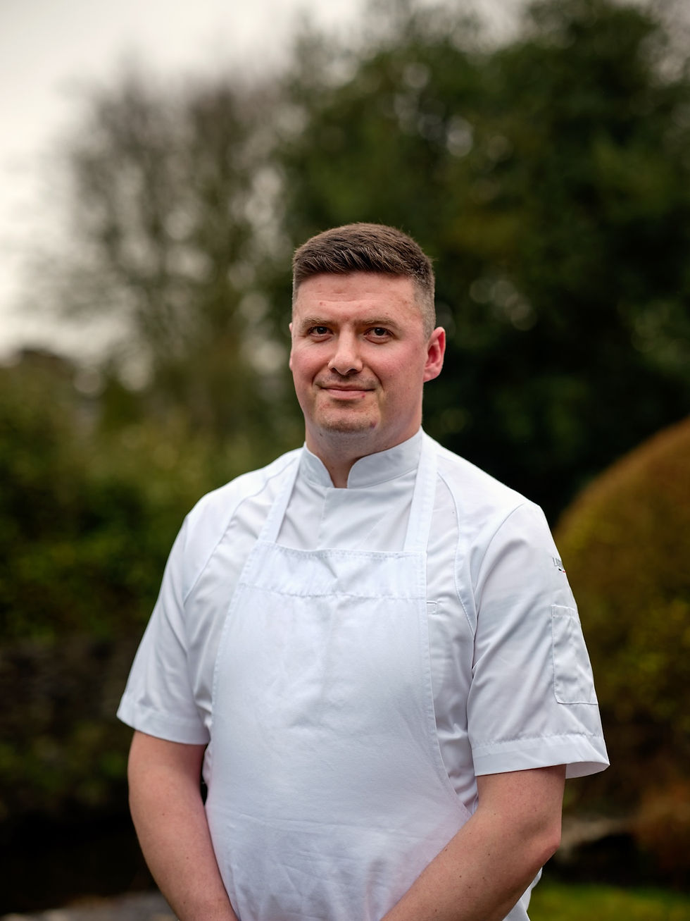 Smiling chef in white coat and apron standing outdoors with blurred green trees in the background.
