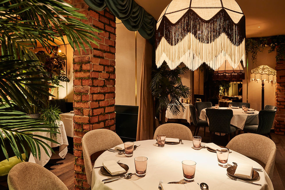 Cozy restaurant with round tables, white cloths, brick walls, palm plants, and large fringed lamps. Warm lighting and elegant setting.