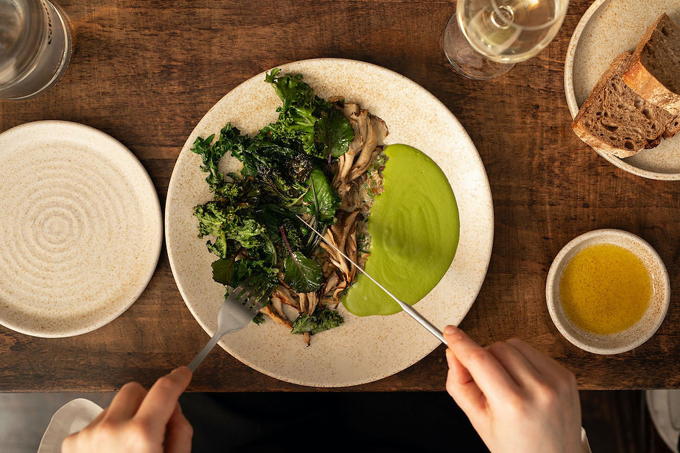 Hands holding cutlery with a dish of greens, mushrooms, and green sauce on a wooden table. Bread, olive oil, and white wine nearby.