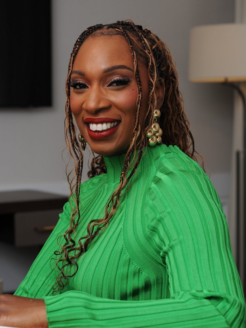 Smiling woman with braided hair in a bright green dress and gold earrings indoors near a lamp.