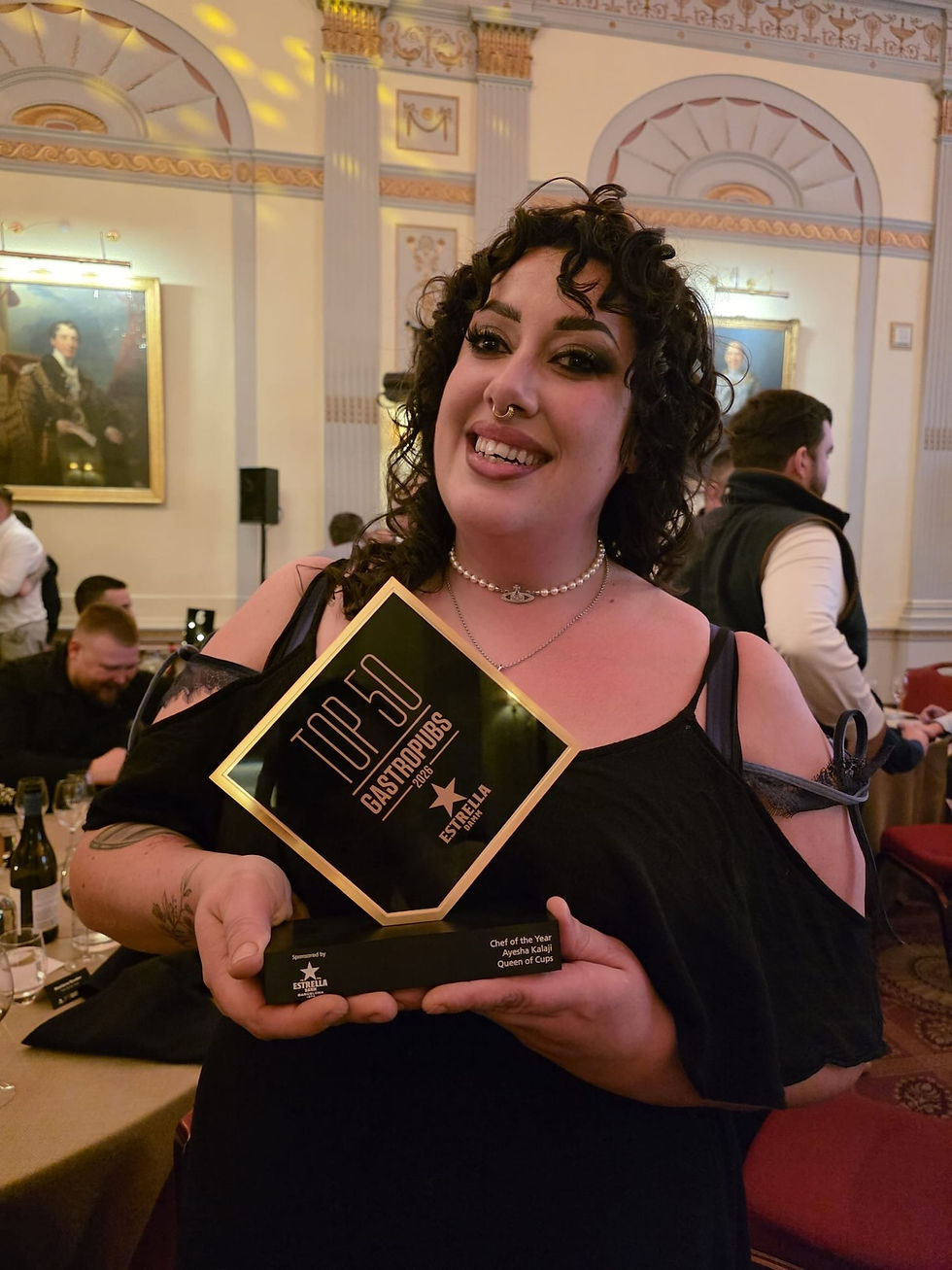 Woman smiling and holding a "Top 50 Gastropubs" award in an elegant room. Background includes ornate decor and a historical painting.