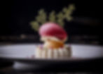 Elegant dessert on a plate: white base, orange sphere, red sorbet, garnished with green leaves and purple flowers, set against dark background.