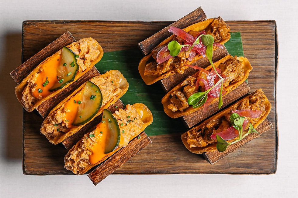 Mini tacos topped with pickled onions, cucumbers, and green garnish on a wooden board with a banana leaf, showcasing vibrant colors.