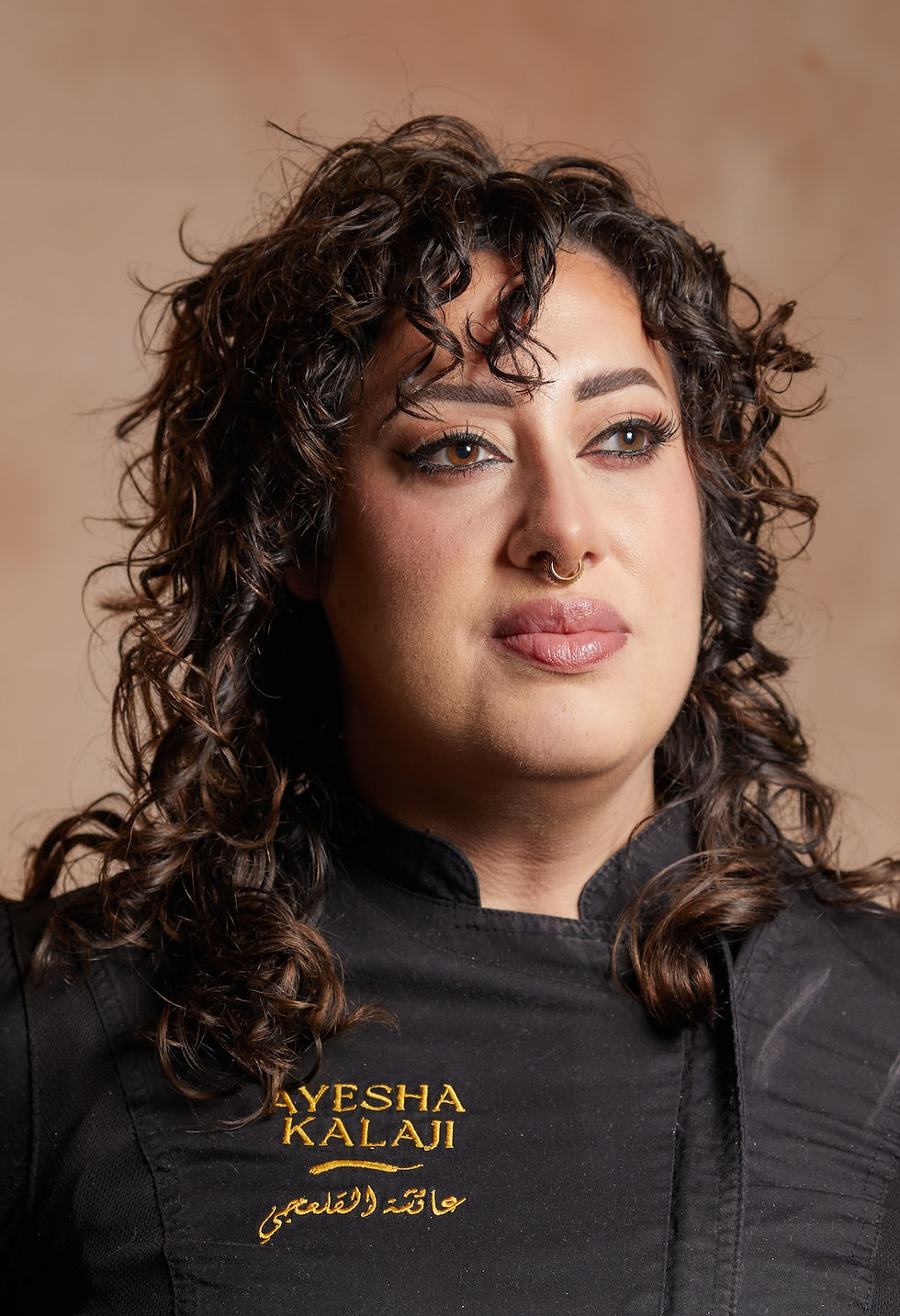 Person with curly hair wearing a black shirt with gold text "AYESHA KALAJI." Neutral background, calm expression, visible nose ring.